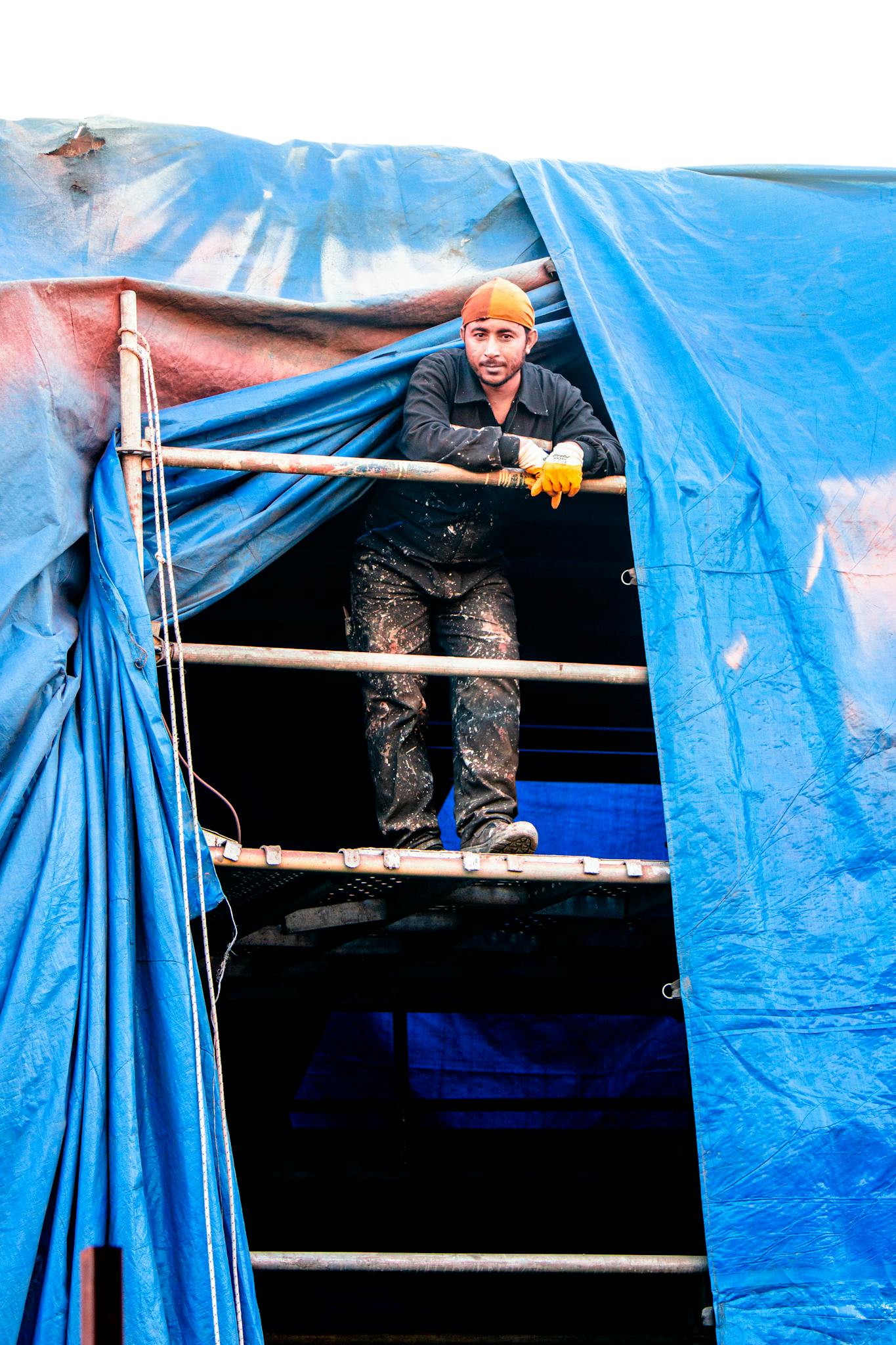 A construction worker stands on scaffolding wrapped in a blue tarp, wearing protective gear.