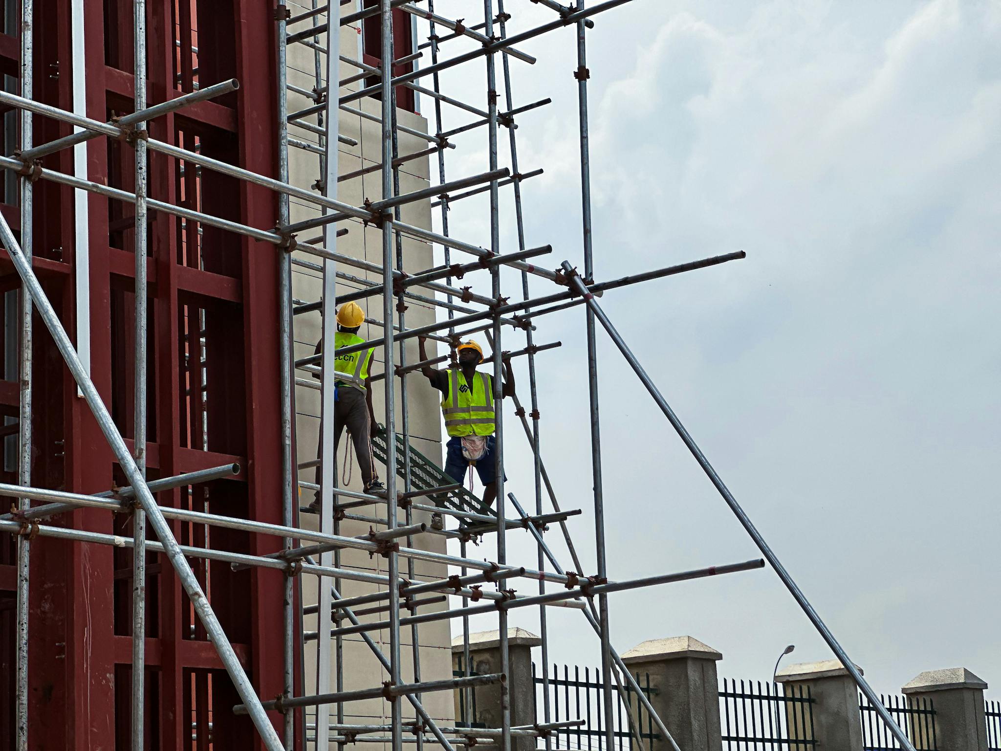 Two workers in safety vests and helmets work on scaffolding, set against a cloudy sky.